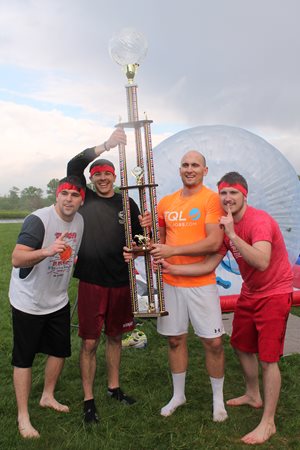 Four Employees Smiling In Front Of An Inflatable Hamster Ball And A Tall Trophy